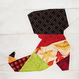 May include: A patchwork quilt block in the shape of a boot, featuring a variety of fabrics in shades of red, green, brown, and yellow. The fabrics have different patterns, including plaid, floral, and geometric designs.