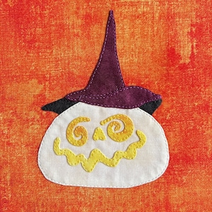 May include: A Halloween-themed fabric applique featuring a smiling pumpkin wearing a purple witch hat. The pumpkin has yellow embroidered eyes and a zig-zag mouth. The background is a textured orange fabric.