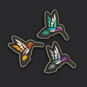 Puede incluir: Tres parches de goma con forma de colibrí y detalles en relieve. Uno presenta acentos en turquesa, blanco y rosa. Otro muestra colores naranja, amarillo y blanco. El tercero exhibe tonos grises, blancos y morados, todos con bordes negros.