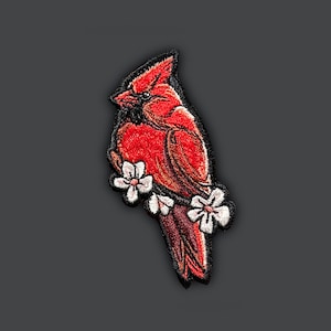 Puede incluir: Un pájaro cardenal rojo con ojos negros y pico negro posado en una rama con flores blancas. El pájaro está bordado con puntadas detalladas.