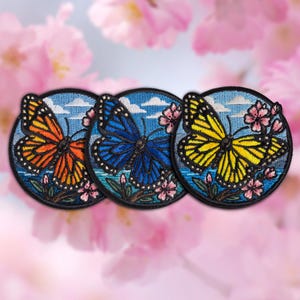 May include: Three embroidered patches featuring butterflies in orange, blue, and yellow. Each patch has a black border, a blue sky background with white clouds, and pink floral accents. The butterflies are the focal point of the design.