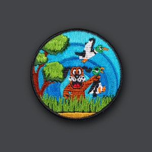May include: Embroidered patch featuring a cartoon dog holding a duck, with a flying duck in the background. The patch is surrounded by green grass and a blue sky with a green tree.