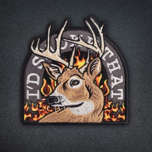 May include: Embroidered patch with a deer head and antlers, encircled by flames. The text "I'D SAY THAT" is in white. The deer is brown and tan, set against a black and grey background. The patch is rectangular.