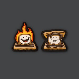 May include: Two embroidered patches depicting a smiling marshmallow being roasted over a fire and a finished s'more with chocolate and graham crackers.