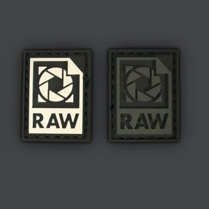 Puede incluir: Dos parches en blanco y negro con un icono de cámara y la palabra "RAW" en ellos. Los parches están cosidos sobre un fondo gris oscuro.