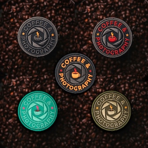 Op de afbeelding: Vijf ronde geborduurde patches met een koffiekopje en een bliksem in een camera lens. De tekst "Coffee & Photography" staat rond de buitenkant van de lens. De patches zijn in verschillende kleuren: zwart, groen, rood en bruin.