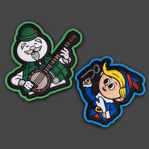 May include: Two cartoon patches. One features a white character with a green hat and plaid vest playing a banjo. The other shows a cartoon elf in blue with a gold buckle and holding a tool. Both have black borders.