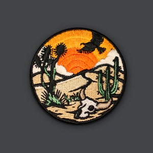 May include: Embroidered patch with a desert scene featuring a sunset, two cacti, a skull, and a flying bird. The patch is circular and has a brown, orange, green, and black color scheme.