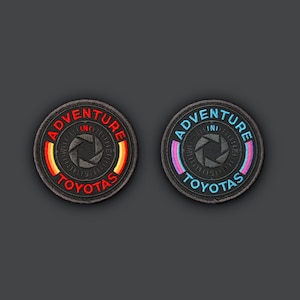 Puede incluir: Dos parches bordados negros con las palabras "Adventure" y "Toyota" en blanco. Un parche tiene una franja roja y naranja, el otro tiene una franja azul y morada.