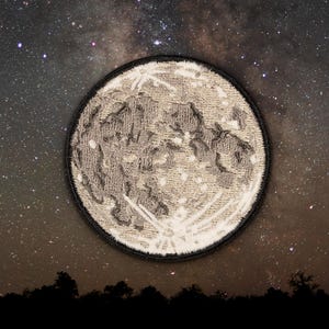 May include: A close-up of a grey and white embroidered moon patch against a dark background of a starry night sky.