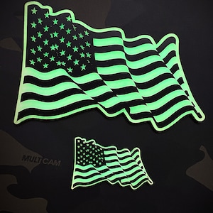 May include: Two glow-in-the-dark American flag patches. The larger patch is waving in the wind. The smaller patch is flat. Both patches are black and green.