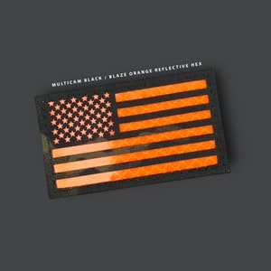 May include: A rectangular patch featuring the American flag design. The stars are orange, and the stripes are blaze orange. The background is a multicam black pattern. The text "MULTICAM BLACK / BLAZE ORANGE REFLECTIVE HEX" is printed above the flag.