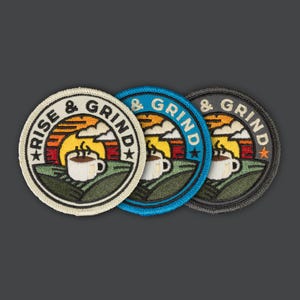 Op de afbeelding: Drie geborduurde patches met een retro zonsopgang ontwerp en een koffiekopje. De patches zijn beige, blauw en zwart. De tekst op de patches luidt "Rise & Grind".