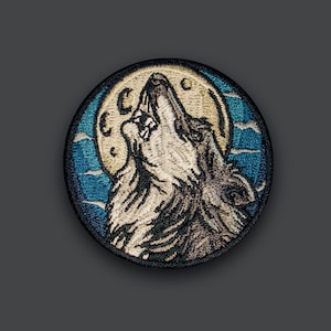 Pode incluir: Etiqueta bordada com um lobo a uivar para a lua. O lobo é retratado em tons de cinza e branco, com um contorno preto. A lua é amarela com um céu azul atrás. A etiqueta é circular e tem uma borda azul e branca.