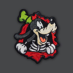 May include: A red, black, and green embroidered patch of a cartoon dog with a skeleton costume peeking out of a torn red background.