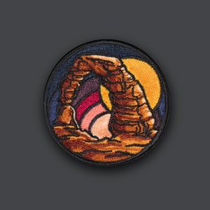 May include: Embroidered patch featuring a sunset-colored arch rock formation against a dark blue background. The arch is brown and orange with a pink and purple sunset behind it.