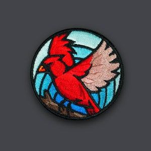 Puede incluir: Un pájaro cardenal rojo con las alas extendidas está bordado en un parche redondo. El fondo es un diseño azul y blanco con una rama marrón.