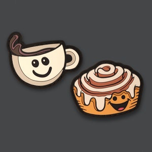 Puede incluir: Dos parches de fieltro, uno de una taza de café marrón con un remolino de crema y una cara sonriente, y el otro de un rollo de canela con glaseado blanco y una cara sonriente.