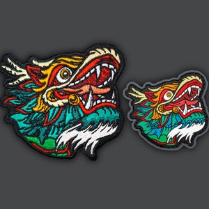 Może przedstawiać: Dwa haftowane naszywki przedstawiające kolorową głowę smoka z otwartą paszczą i ostrymi zębami. Większa naszywka jest bardziej szczegółowa, z zielonym, niebieskim i białym tłem. Mniejsza naszywka jest prostsza, z czarnym tłem.