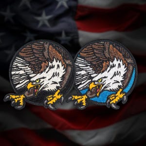 May include: Two embroidered patches featuring a detailed eagle design. The eagles have brown and white feathers, yellow beaks and talons, and are set against a black background. One patch has a blue background. The patches are circular.
