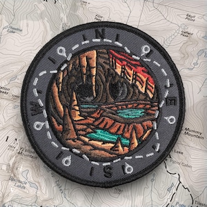 May include: A round, embroidered patch with a landscape design. The patch features a cave scene with a turquoise lake, brown rock formations, and a red and orange sunset. The text "LIMITLESS" is embroidered around the edge in white.