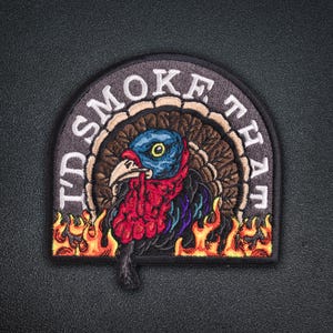 May include: Embroidered patch featuring a turkey with a blue head, red wattle, and colorful feathers, surrounded by flames. The patch has the text "I'D SMOKE THAT" in white lettering on a gray background. A fun, humorous design.