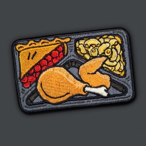 May include: A gray and black embroidered patch depicting a Thanksgiving dinner meal. The patch features a pie, a chicken leg, and a side dish of mashed potatoes.