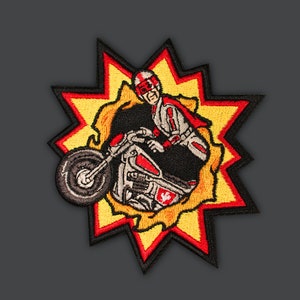 May include: A red and yellow embroidered patch featuring a cartoon character riding a motorcycle through a fiery explosion. The character is wearing a red helmet and a red and white suit.