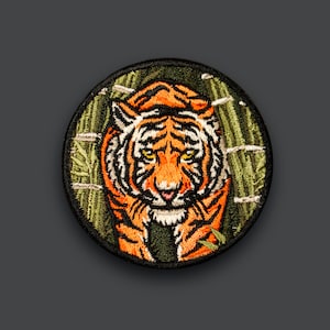 Op de afbeelding: Ronde geborduurde patch met een tijgerontwerp. De tijger heeft oranje en zwarte strepen, gele ogen en een witte snuit. De patch heeft een zwarte rand en een groene bamboe achtergrond.
