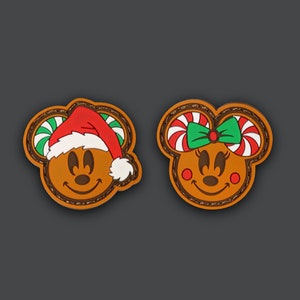 Può includere: Due decorazioni a forma di biscotto di pan di zenzero, una con un cappello di Babbo Natale rosso e l'altra con un fiocco di bastoncino di zucchero verde e rosso. Entrambe hanno occhi marroni, un naso marrone e una bocca sorridente.