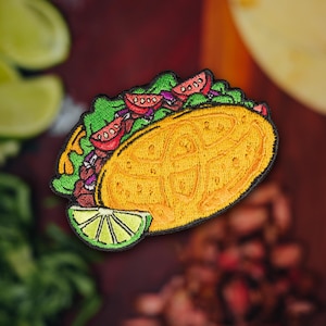 May include: A yellow embroidered taco with red tomatoes, purple onions, and green lettuce. The taco is on a brown background with a lime wedge.