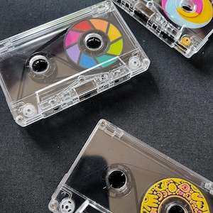 May include: Transparent cassette tapes with vibrant, abstract reel designs. One tape has a rainbow wheel, another a spiral of pink, blue, and yellow, and a third with a yellow design. The tapes are displayed on a dark surface.