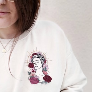 Peut inclure: Un sweat-shirt blanc avec un motif brodé représentant un visage de femme entouré de fleurs roses et rouges. Le motif est de style vintage.