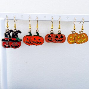 May include: A collection of Halloween-themed earrings. The earrings feature various designs, including jack-o'-lanterns and witch hats. The earrings are made of metal with gold-colored hooks and are displayed on a white earring holder.