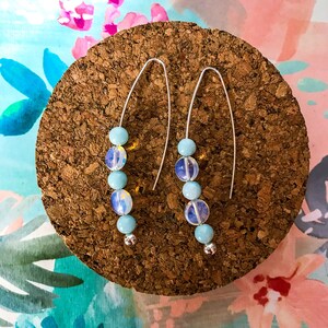 May include: A pair of dangle earrings featuring a silver wire design. Each earring has a series of light blue and iridescent beads, with a small silver bead at the bottom. The earrings are displayed on a cork surface against a colorful floral background.