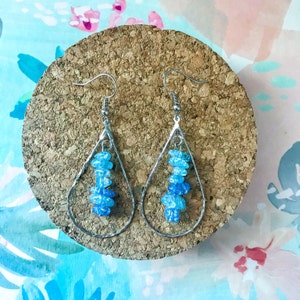 May include: A pair of silver teardrop earrings with blue and light blue glass beads. The earrings are hanging from a cork surface.