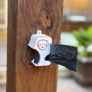 Op de afbeelding: Een witte hondenpoepzakjesdispenser met een rode omtrek en een honden gezichtsgrafiek is op een houten paal gemonteerd. Een zwarte afvalzak wordt uit de dispenser getrokken. De dispenser is ontworpen voor huisdiereigenaren.