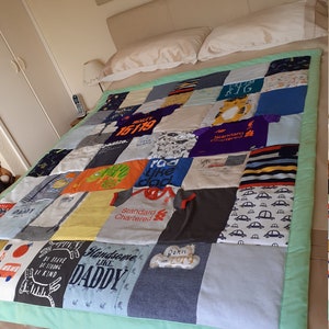 May include: A patchwork quilt made from baby clothes with a green border. The quilt features various designs and text, including "Dream Big", "Standard Chartered", "Born in 2019", and "Handsome Like Daddy".