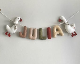 May include: A decorative garland featuring the name JULIA spelled out in plush, stuffed fabric letters in shades of terracotta, beige, olive green, and rust, flanked by two white felt geese with orange beaks and feet, all strung on a white cord.