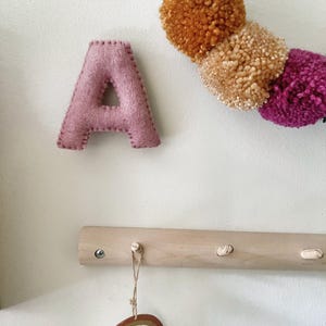 May include: A soft, felted letter A in dusty pink with visible stitching along the edges, positioned near a decorative garland of mustard, tan, and magenta yarn pom-poms and a wooden wall hook rack featuring a hanging rainbow-shaped ornament.