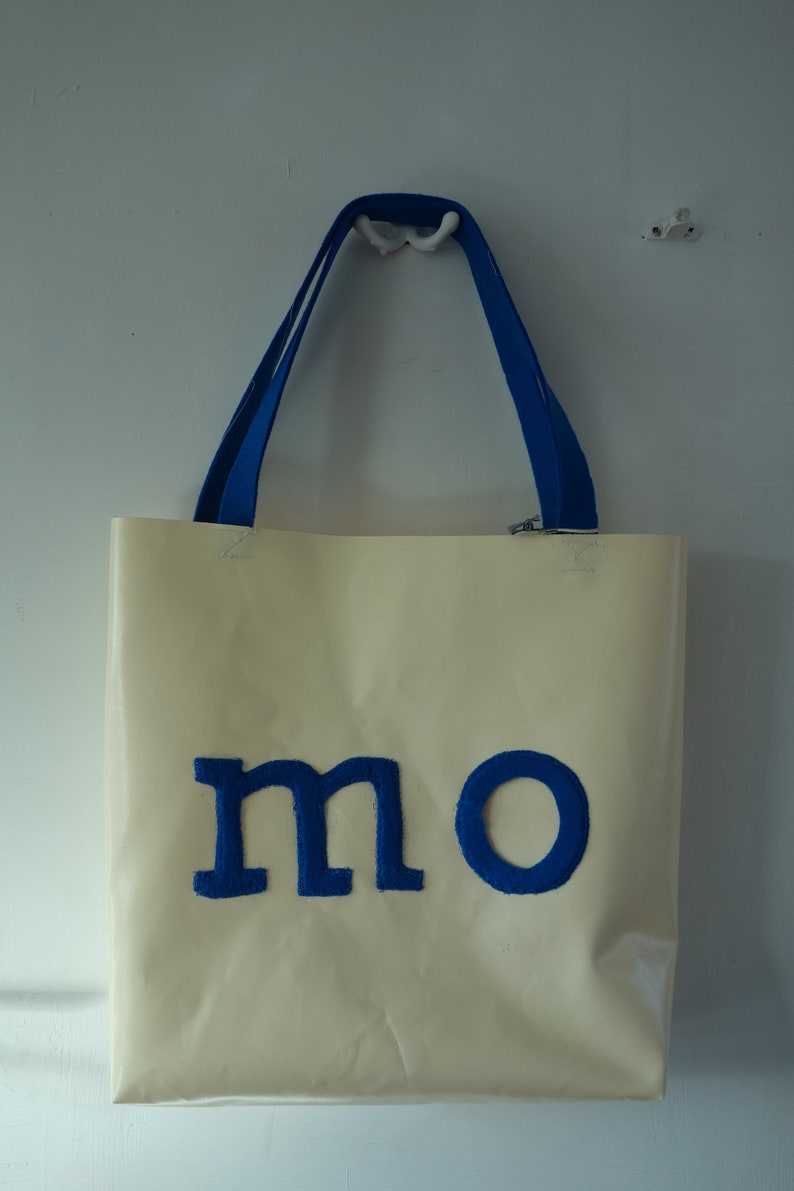 May include: A cream-coloured tote bag with blue handles and the word "mo" in blue felt letters.
