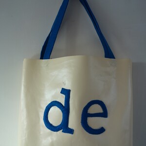 May include: A white tote bag with blue fabric handles and the word "de" embroidered in blue on the front.