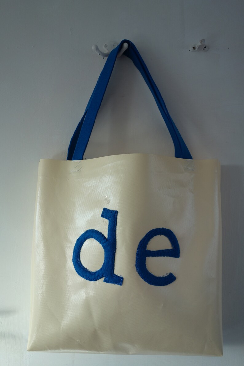 May include: A white tote bag with blue fabric handles and the word "de" embroidered in blue on the front.