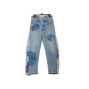 May include: Light wash denim jeans with blue floral designs and patchwork accents. The jeans are hanging on a black hanger. The patchwork is on the outer seams of the jeans.