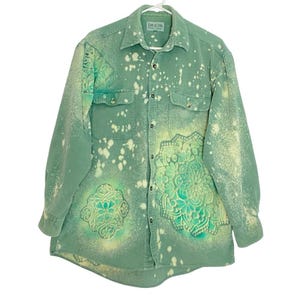 May include: A green button-down shirt with a unique design. The shirt features a bleached effect with white splatters and two turquoise mandala designs. The shirt has two chest pockets and is hanging on a white hanger.