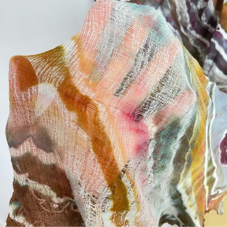 May include: A close-up of a sheer, white fabric with a hand-dyed pattern in shades of brown, pink, and green. The fabric has a loose weave and a slightly textured surface.