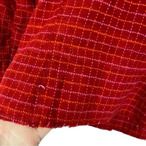 May include: Red fabric with a pink and orange grid pattern. The fabric has a frayed edge.