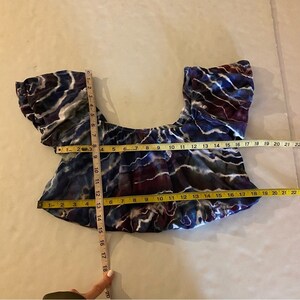 May include: A blue, purple, and white tie-dye off-the-shoulder crop top with short puff sleeves. The top is measured with a tape measure.