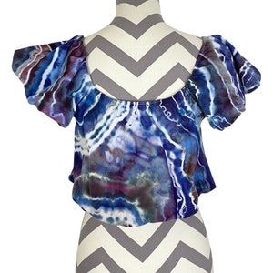 May include: A blue, purple, and white tie-dye off-the-shoulder crop top with puffed sleeves. The top is made of a soft, stretchy fabric and has a relaxed fit.