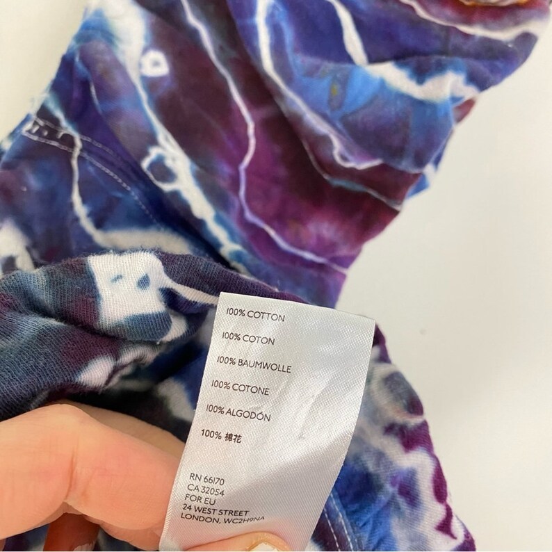 May include: A white tag with black text on a tie-dye fabric background. The tag lists the fabric content as 100% cotton in English, French, German, Italian, Spanish, and Chinese. The tag also includes the company's contact information: RN 66170, CA 32054, FOR EU, 24 WEST STREET, LONDON, WC2H9NA.